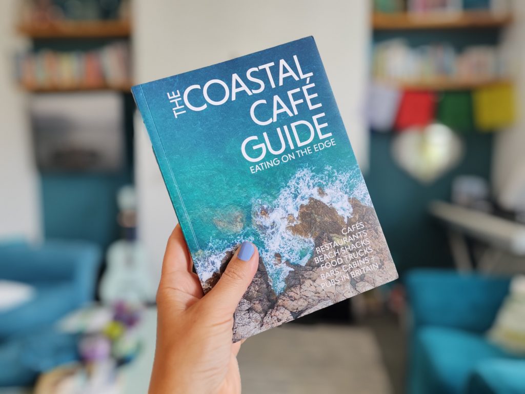 Hand holding The Coastal Café Guide in a living room.